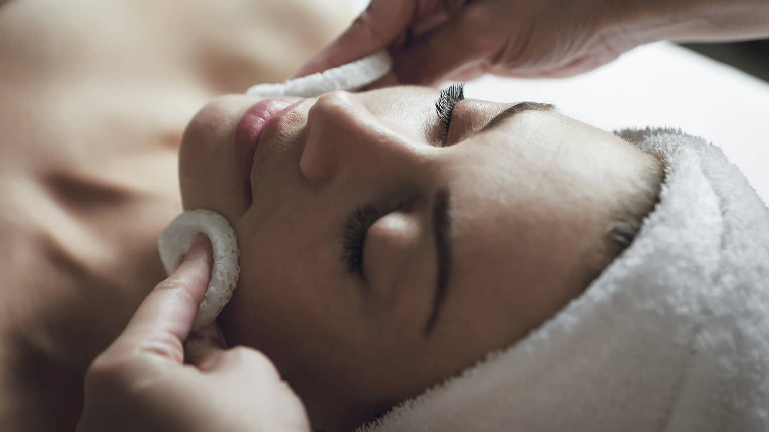 A detail of hands rubbing a sponge on a woman's face as she lies on a massage table with her eyes closed and hair wrapped up in a white towel