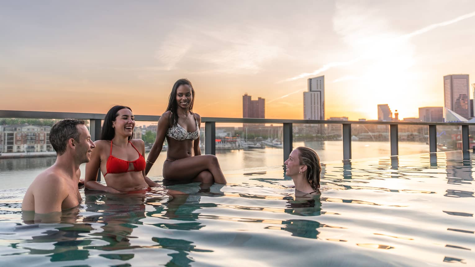 Four people relax by the edge of a swimming pool with the sun setting over the city skyline behind them