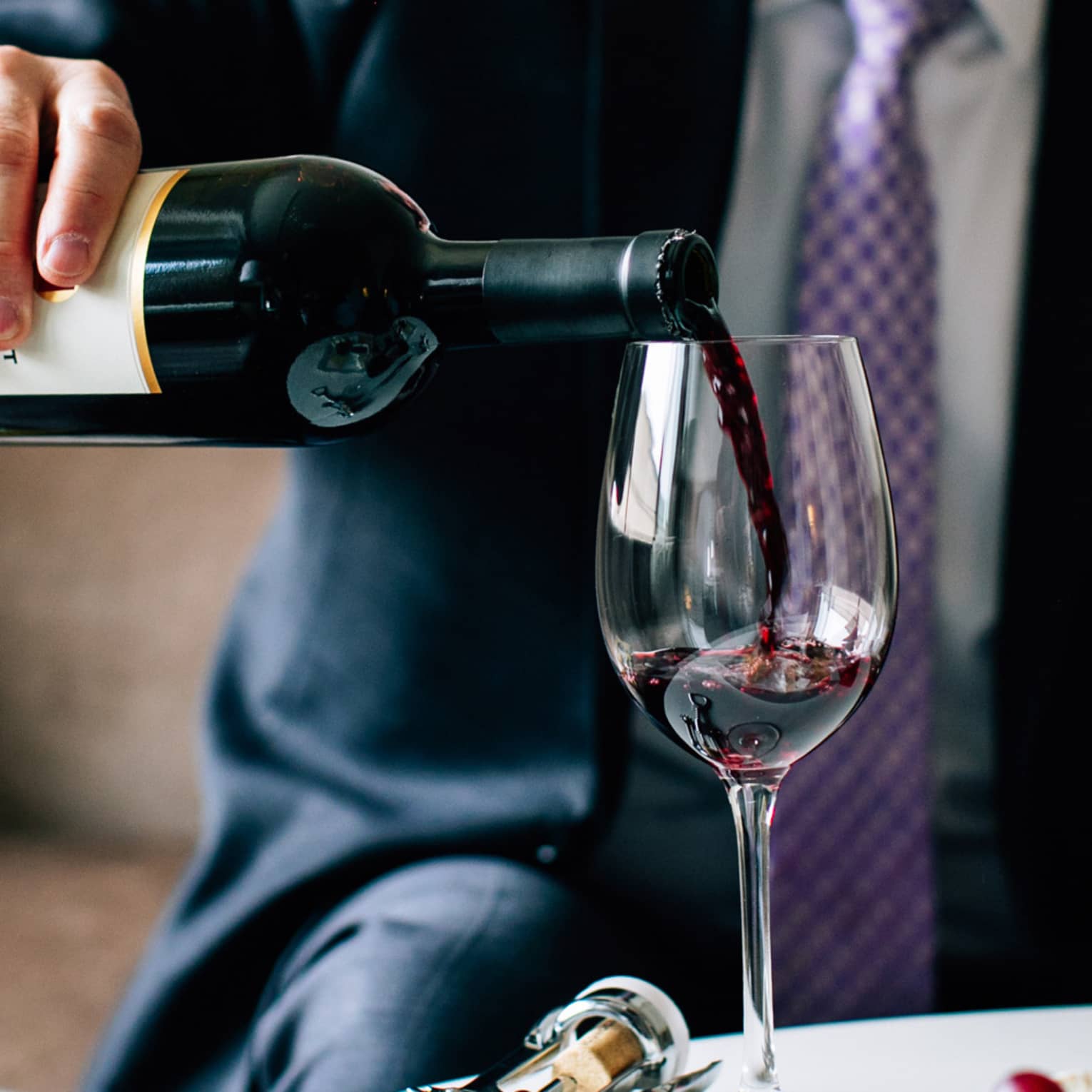 Close-up view of poured red wine cascading into a glass, held by a person with a jacket and tie.
