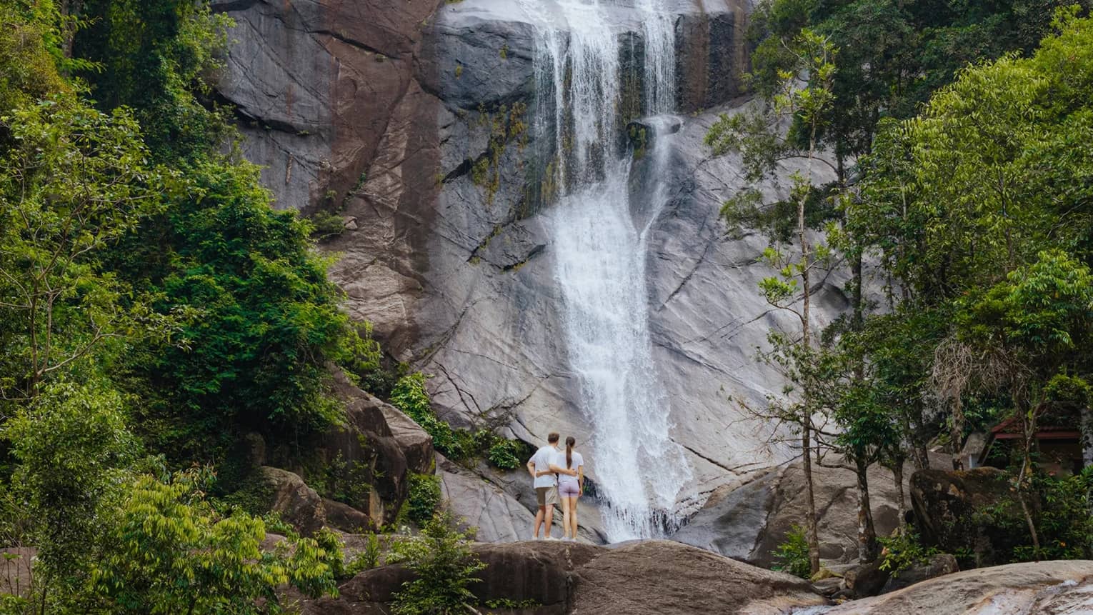 A couple stands at the base of a waterfall surrounded by lush green trees