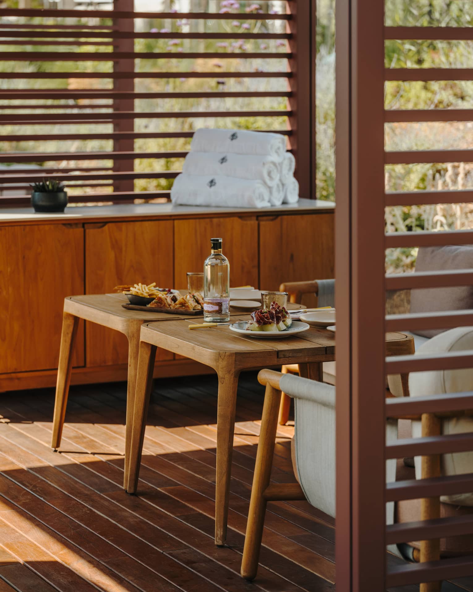 A private pool cabana with small tables, comfortable seating, a cabinet with rolled-up towels and slatted walls admitting filtered light sunlight.