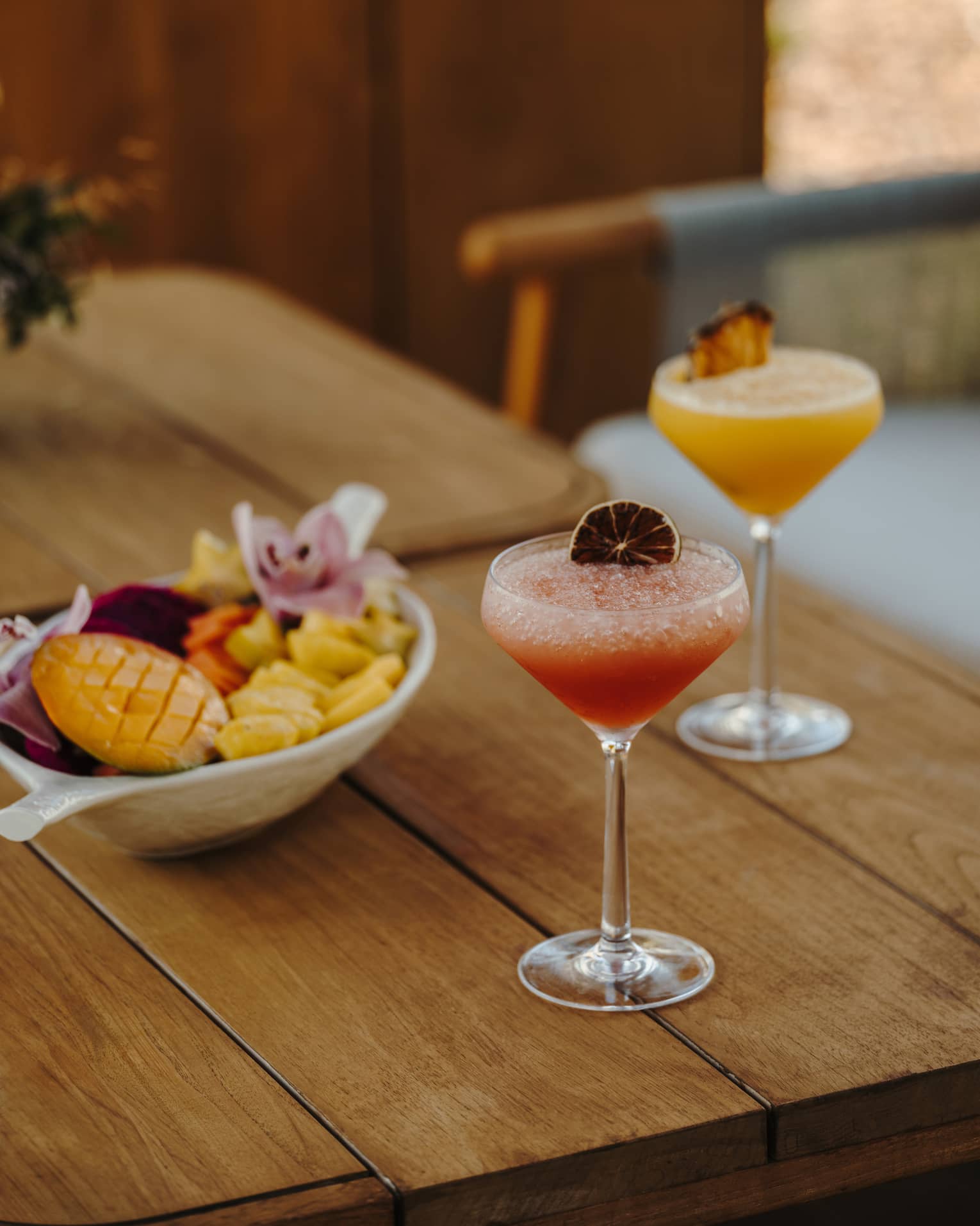 A wood table holds two stemmed glasses of pink and orange drinks with garnishes and a bowl of small-bite food items.