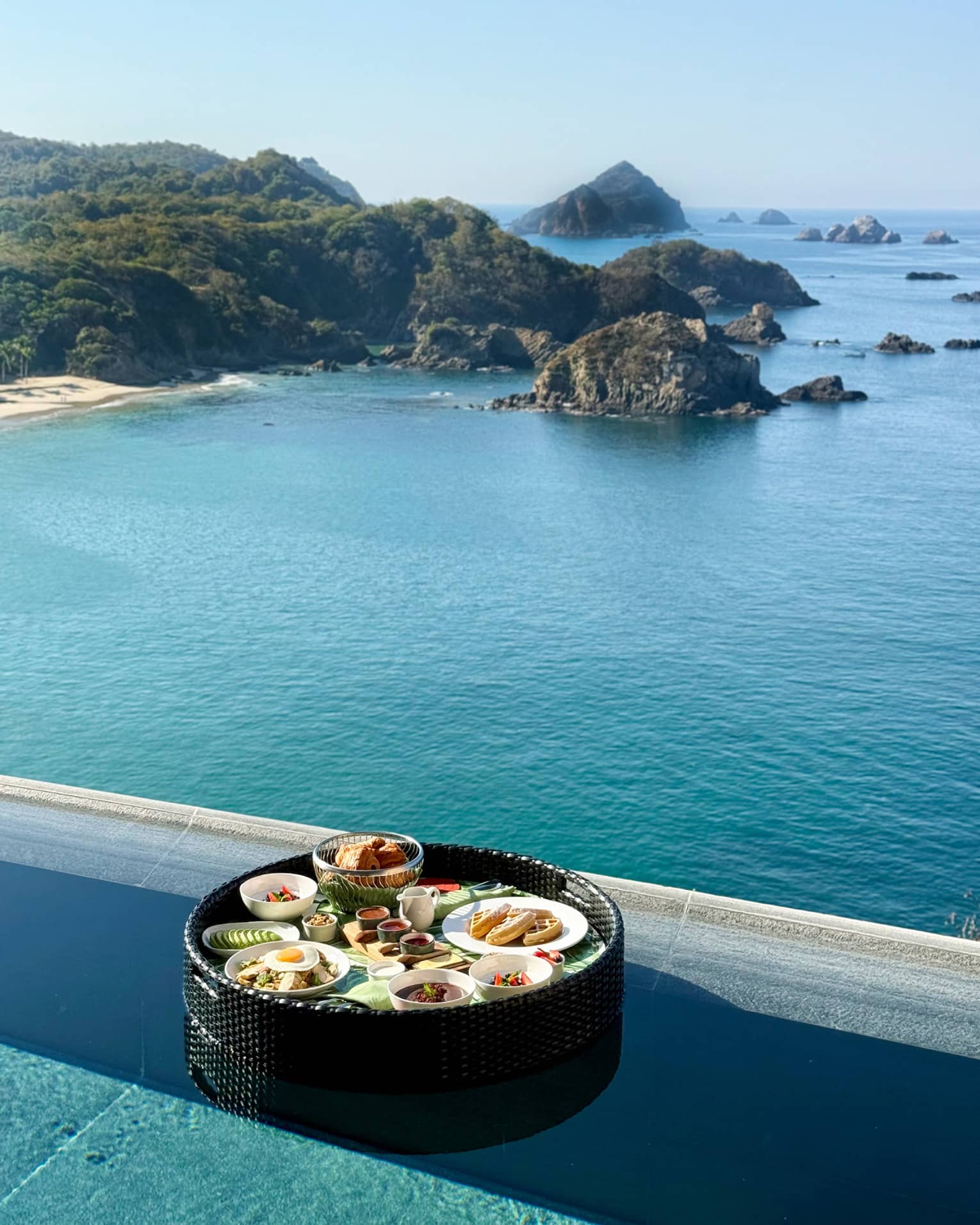 A tray of plated breakfast food items on a tray floating near the edge of an infinity pool, with coastline and ocean views.