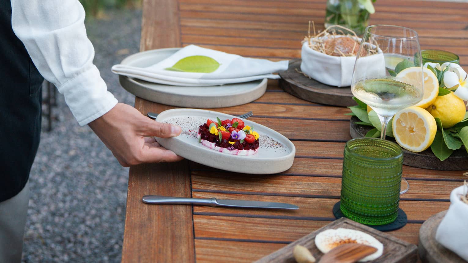 Person setting a dish on a wooden table, with a place setting and wine glass.