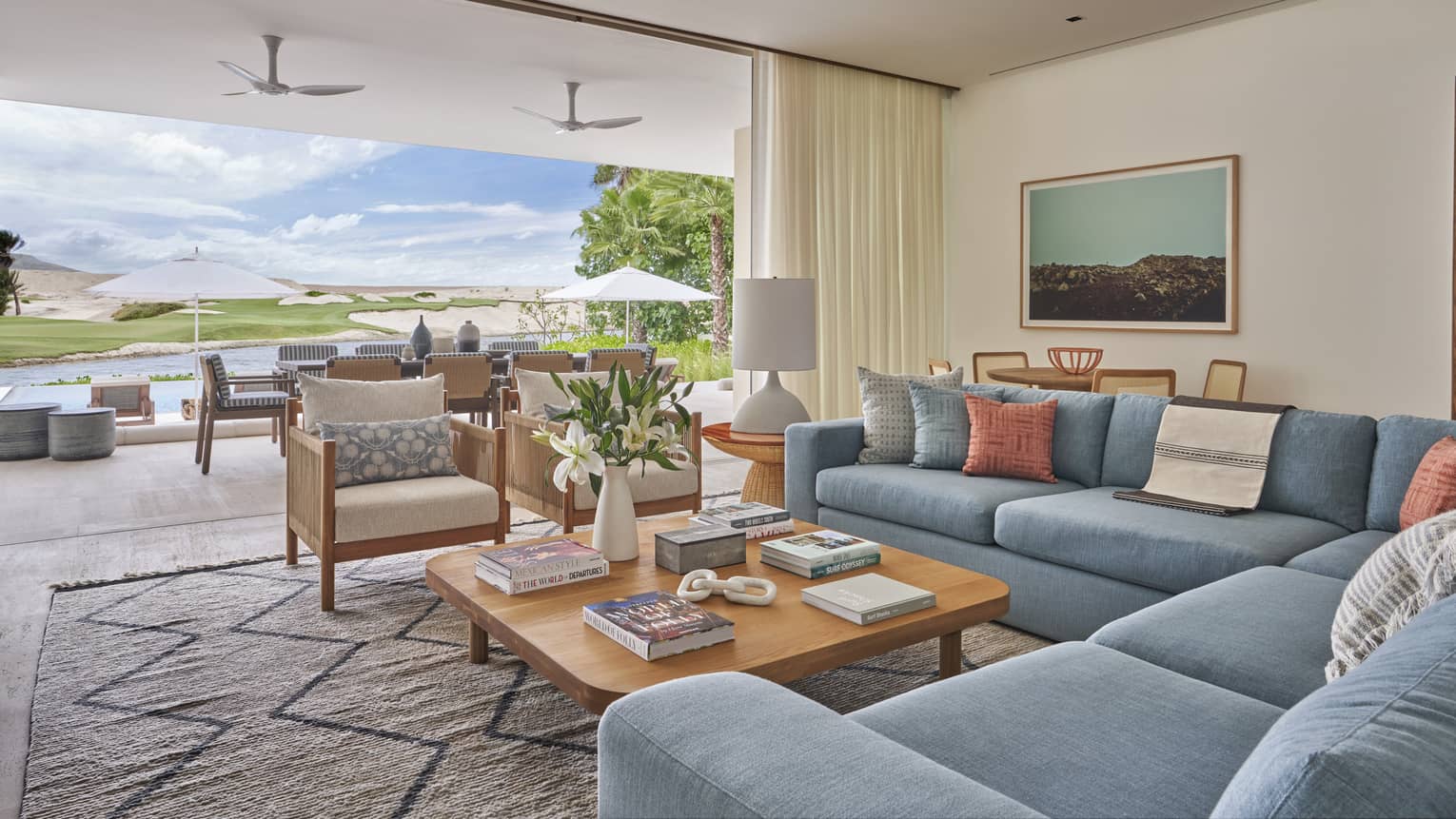 Large modern living room with blue sectional, square wooden coffee table and two club chairs with wall open to terrace and golf course view 