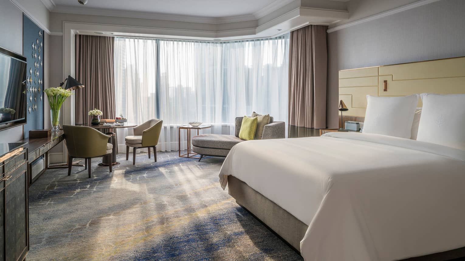 Sunny Deluxe Room bed with white linens, small dining table and chaise lounge, white curtains