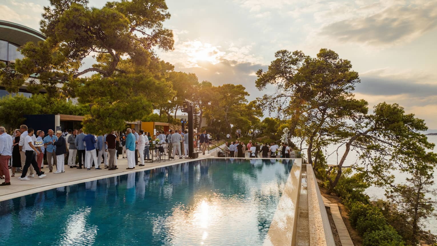 Group of people standing beside infinity pool with view of Saronic Gulf at sunset