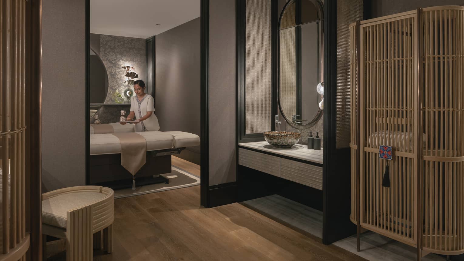 A tray is gently placed on one of two massage tables in a warmly lit spa room with soft grey walls and natural wood décor