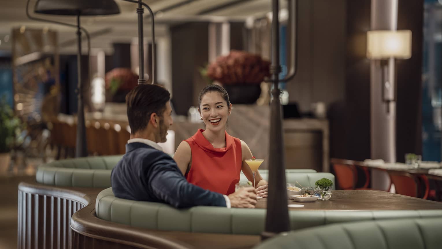 A smiling couple sit at a circular booth in an empty, dimly lit restaurant with cocktails and appetizers on their table.