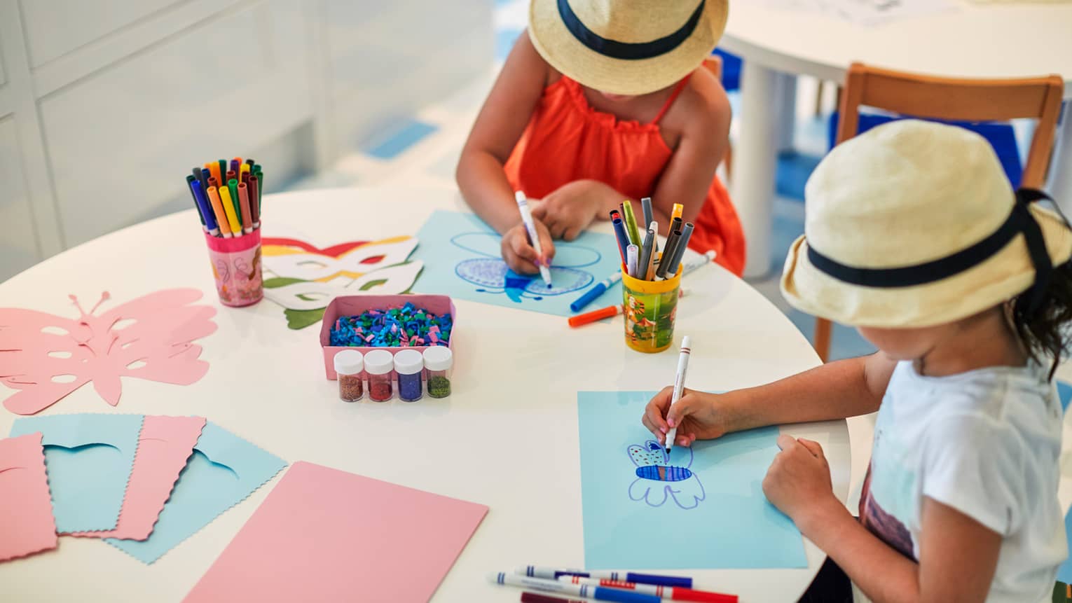 Two young girls wearing straw sun hats colour pictures at craft tables