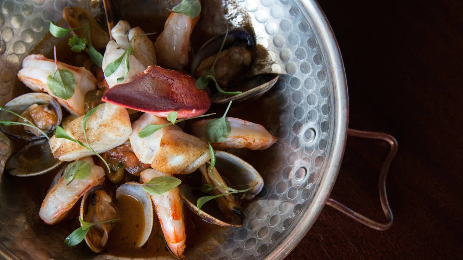 Close-up of a patterned metal bowl filled with prawns, scallops and mussels in a rich, dark broth garnished with green herbs.