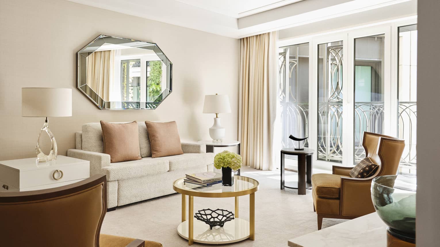 Luxury hotel suite with a neutral sofa, gold-accented round coffee table, brown armchairs, geometric mirror and floor-to-ceiling windows with ornate ironwork