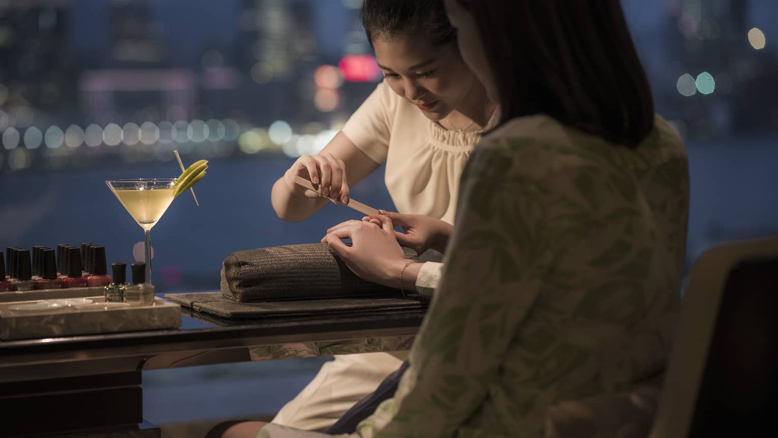 A person gets a manicure at a spa at night, overlooking a harbor. A martini sits on the table beside their hand, with nail polishes in the background.