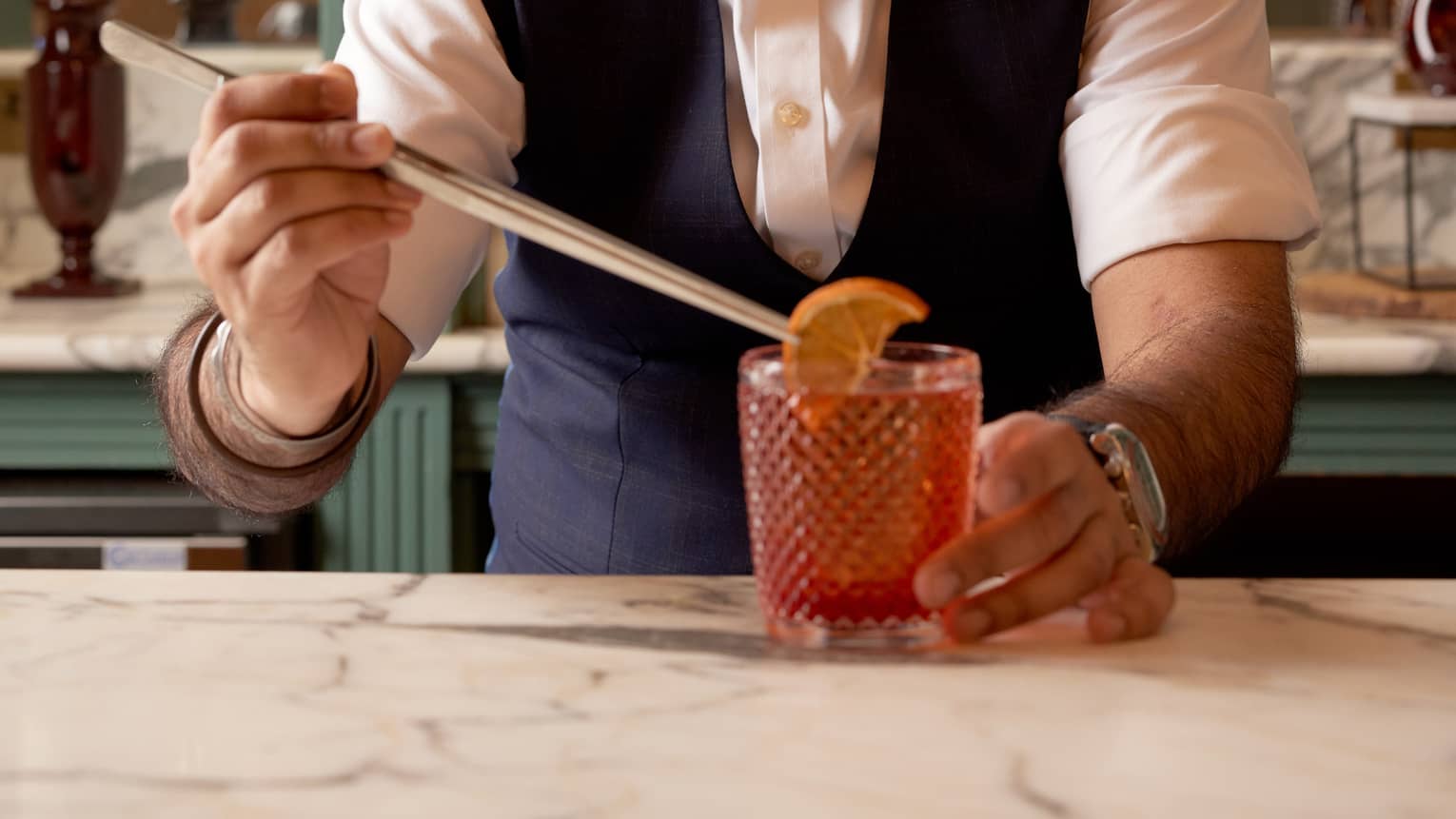 Bartender behind bar garnishing a negroni with an orange slice using long tongs