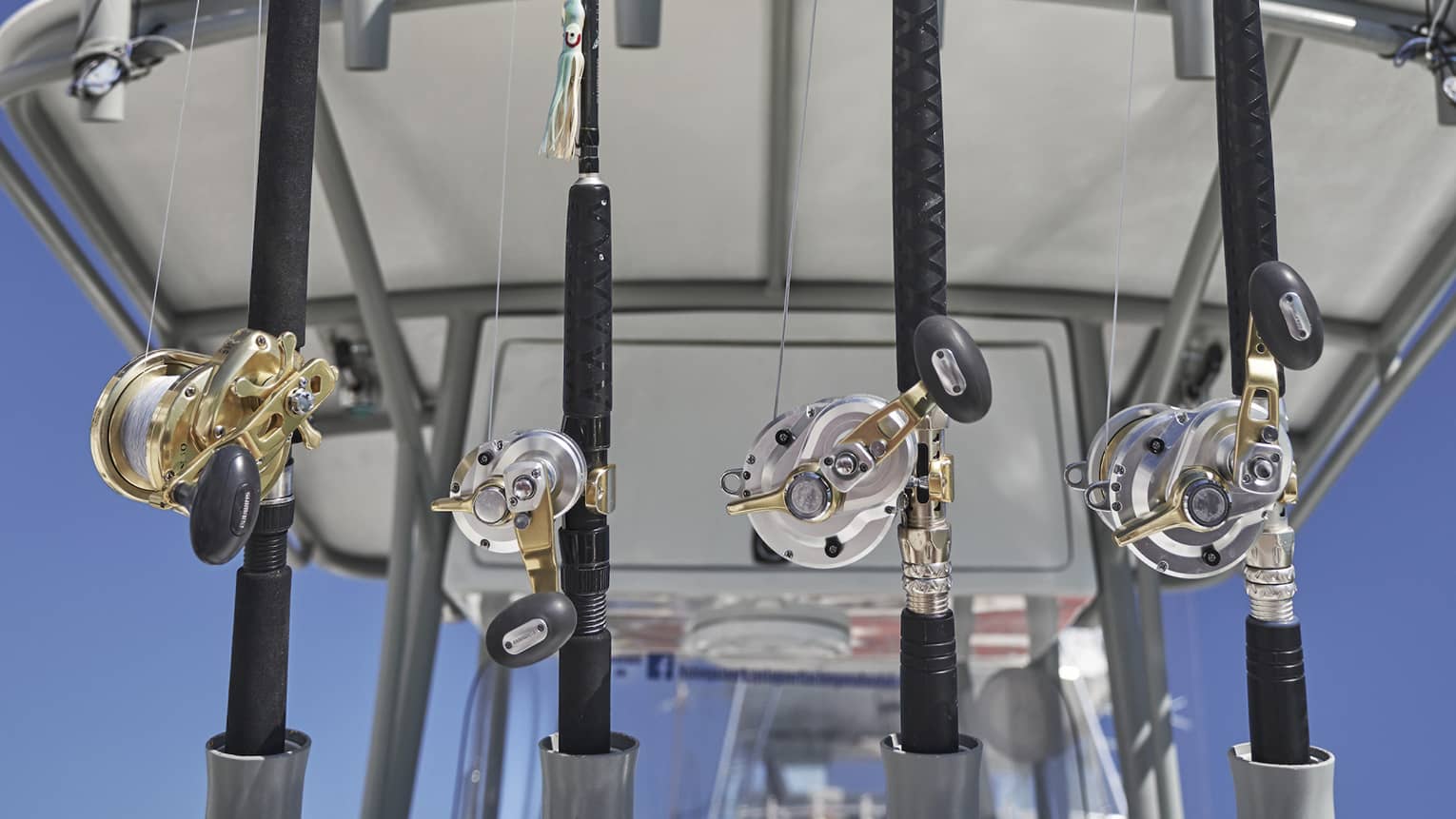 Close-up shot of ten upright fishing rods in a rack attached to a boat’s Bimini top, a clear, dark blue sky behind them.