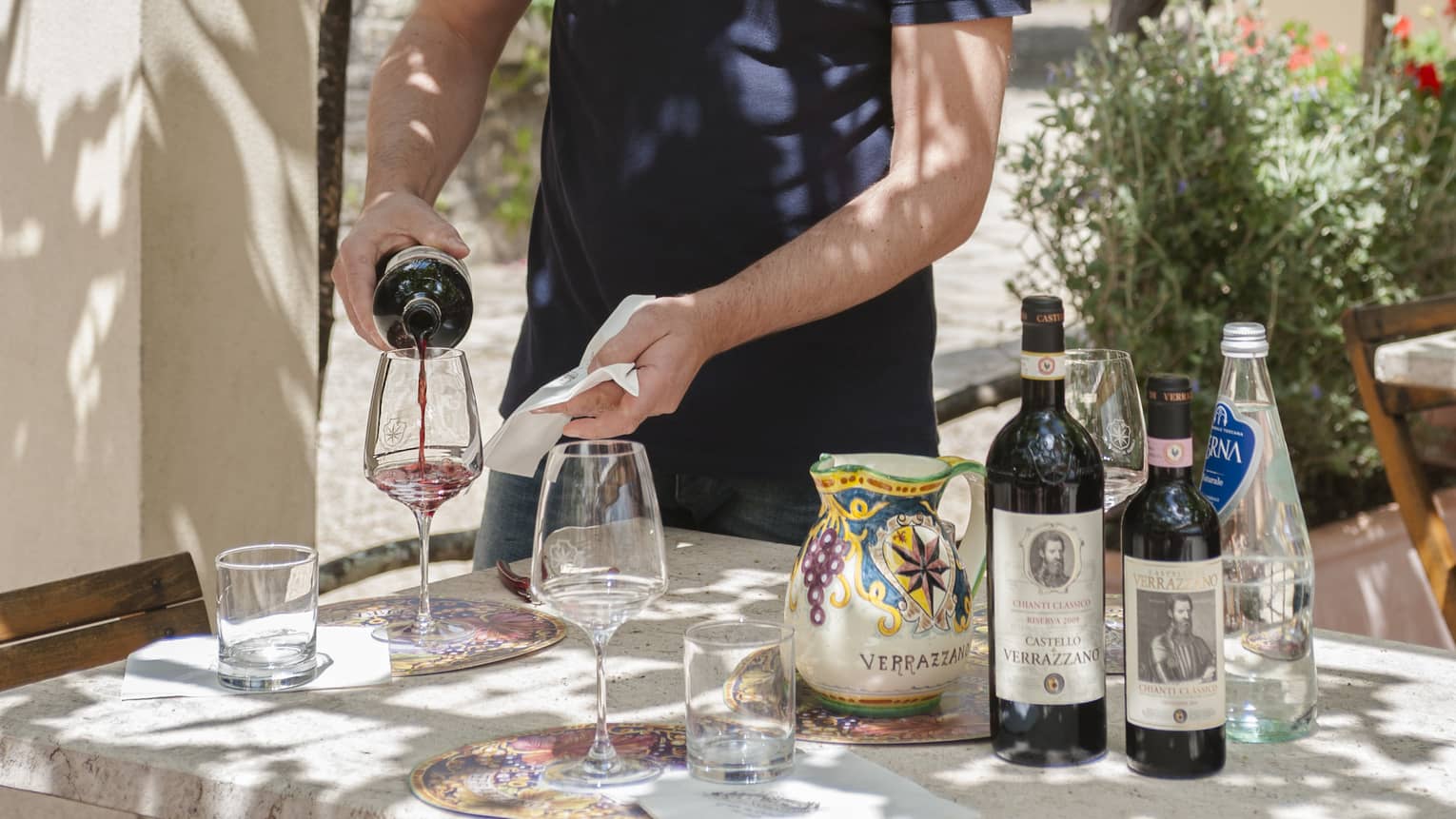 Man wearing apron pours wine at table in sunny yard at Vincigliata Castle 