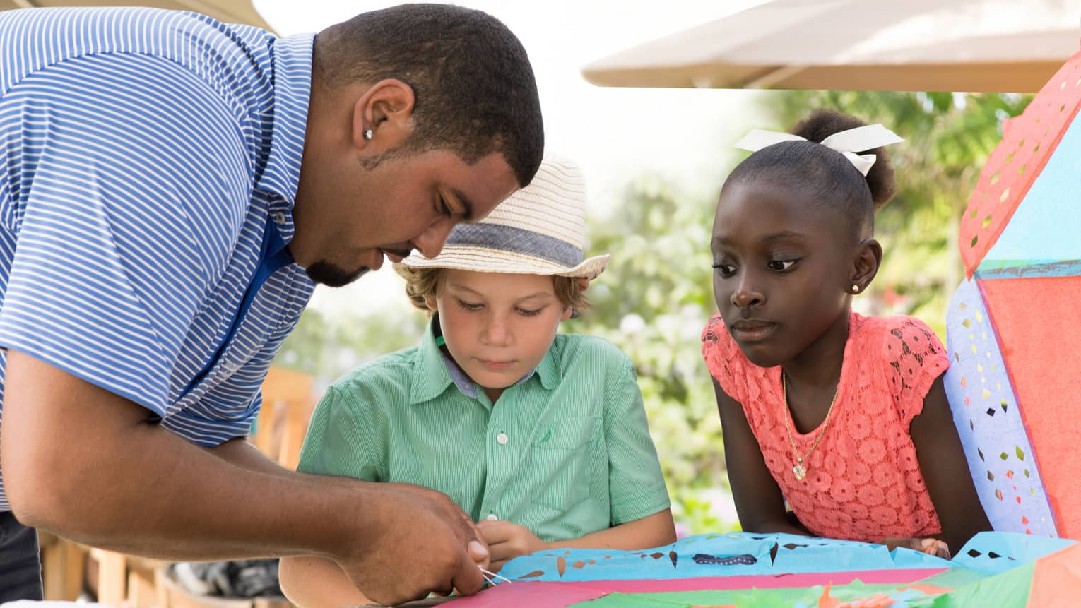 Man ties string onto colourful paper kite at table as two children watch