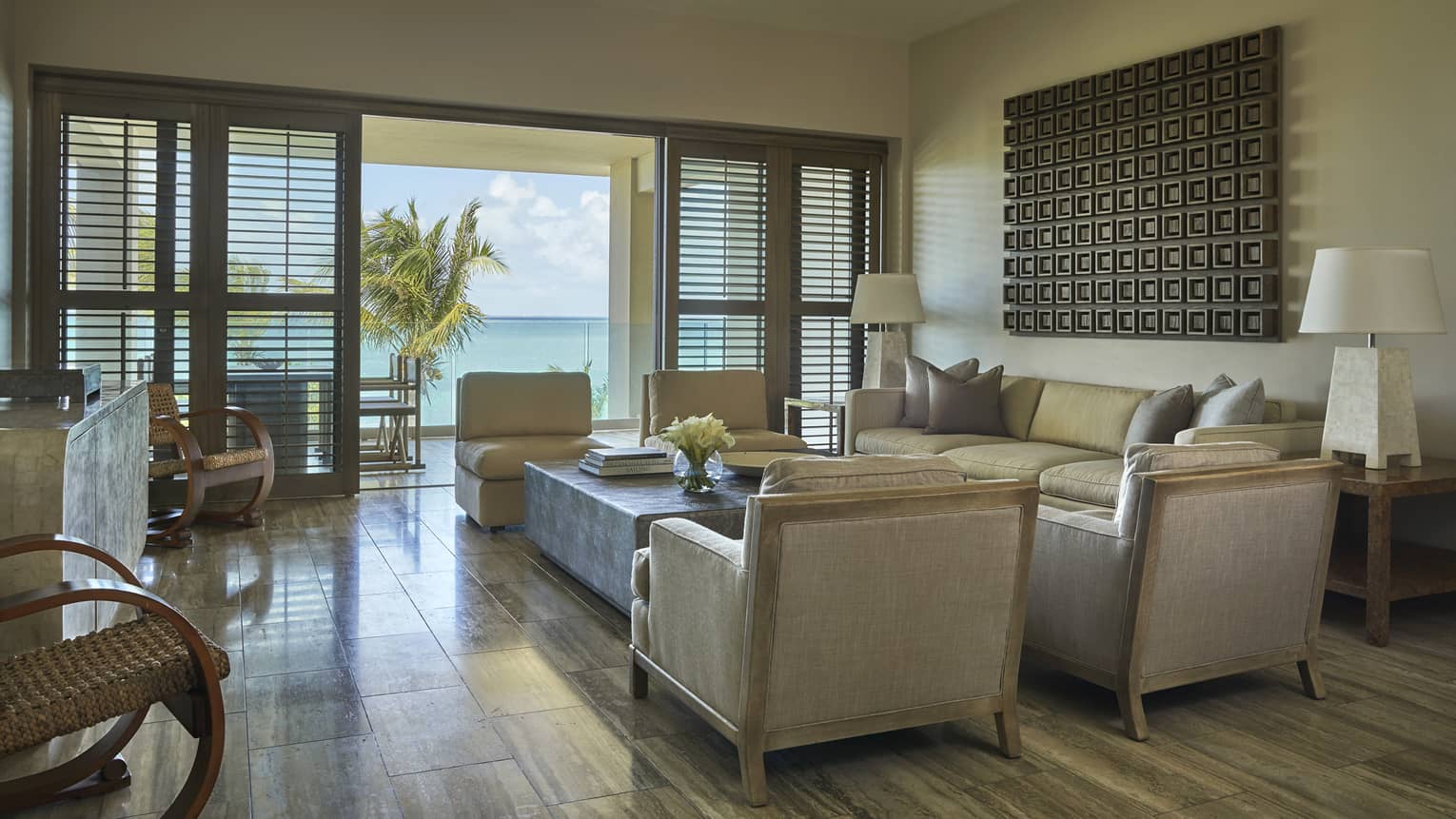 Elegant living area of a resort suite with neutral-toned furniture, modern decor and doors opening to a balcony with ocean and palm tree views