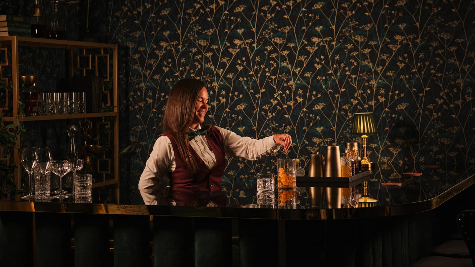 Bartender wearing white shirt, burgundy vest and black bowtie pours a cocktail from behind the bar