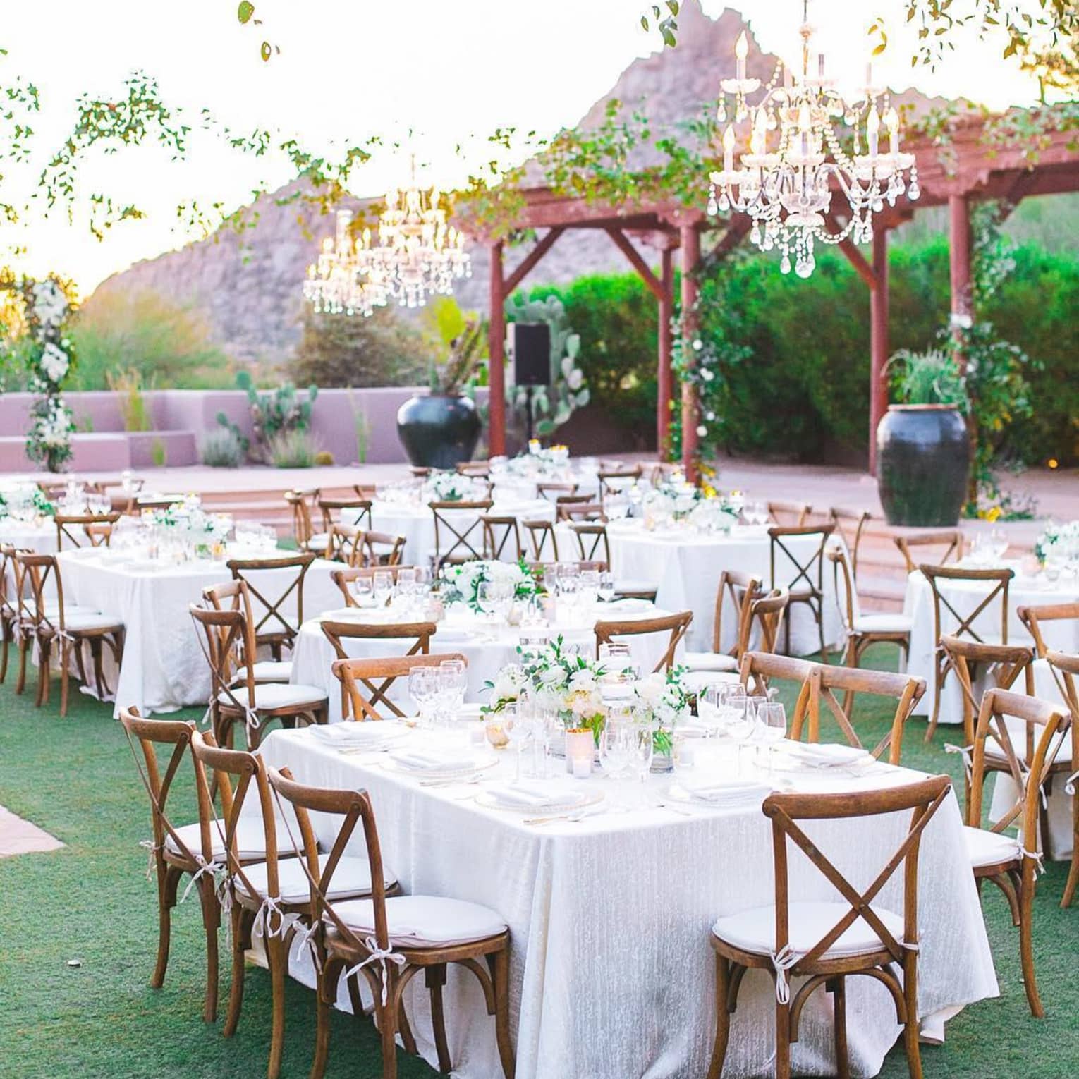 An outdoor wedding reception in a green space with wooden arches, banquet tables decorated with white linens and floral arrangements and greenery and chandeliers hanging above