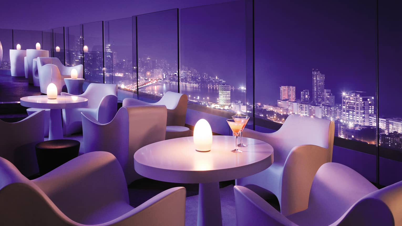 Modern white cocktail tables with candles in lounge with purple lighting, city night views