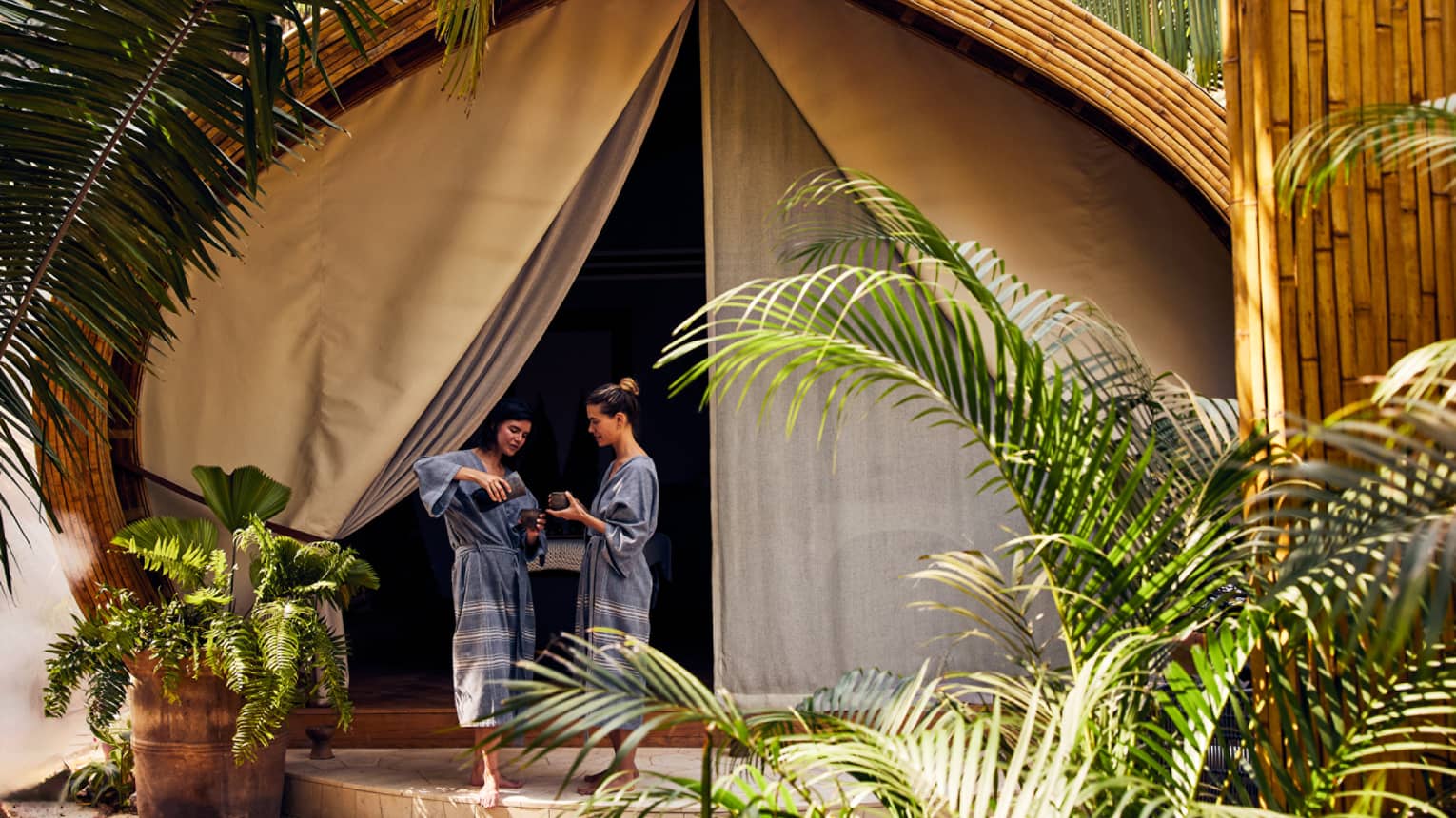 Two women in spa robes stand outside a luxury spa tent, one pouring tea for the other, surrounded by lush greenery in a peaceful setting.