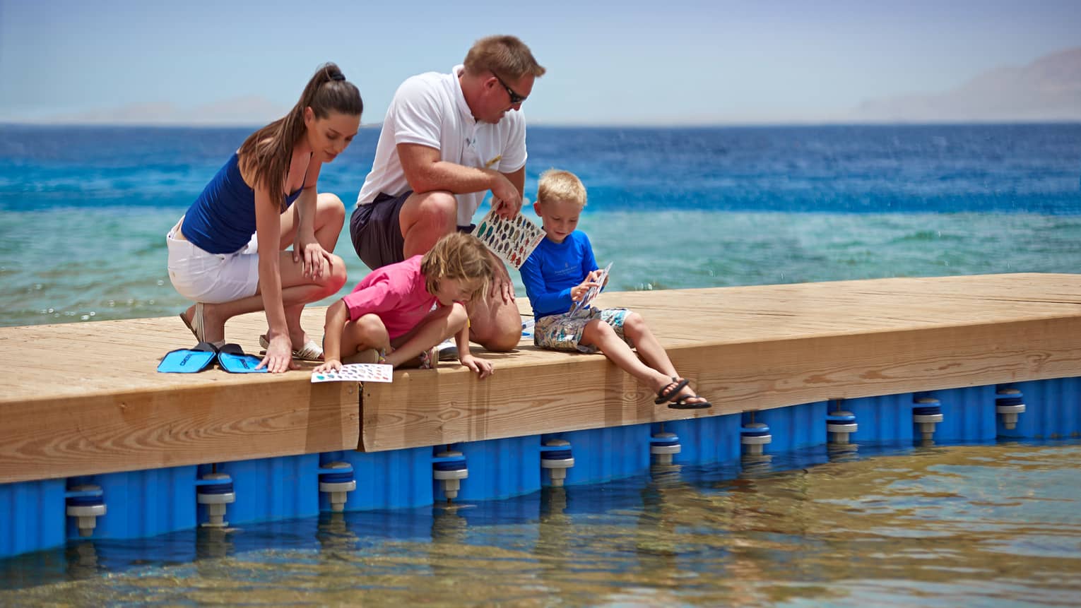 Family holding papers with colourful fish kneel on dock, peer into water