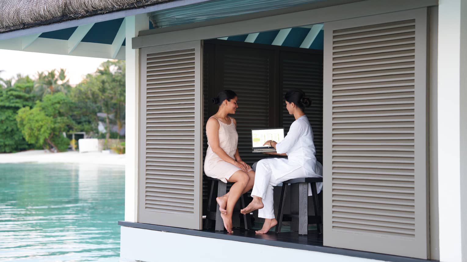 A doctor and a guest sit at a poolside cabana looking over some things on a laptop