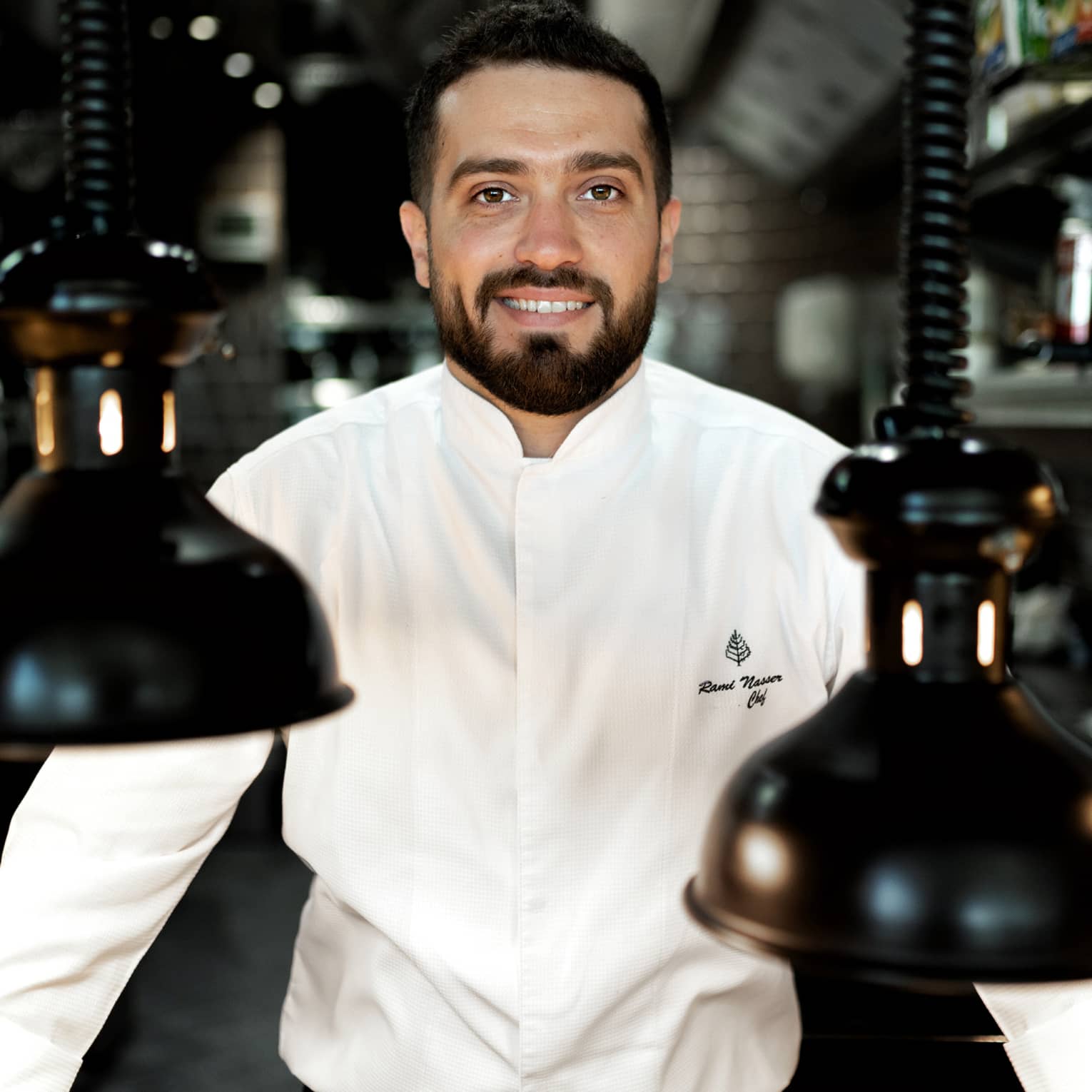Chef Rami Nasser in his white chef's jacket smiles at the camera, flanked by black vintage pendant lamps hanging from coils.