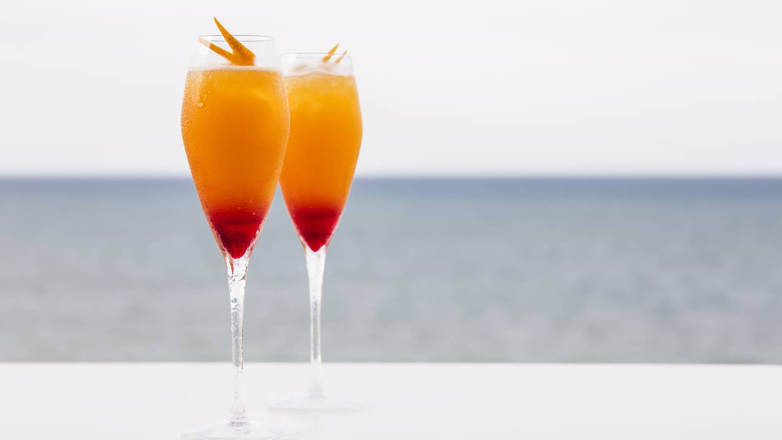 Two cocktails with garnish on table outdoors with ocean in background 