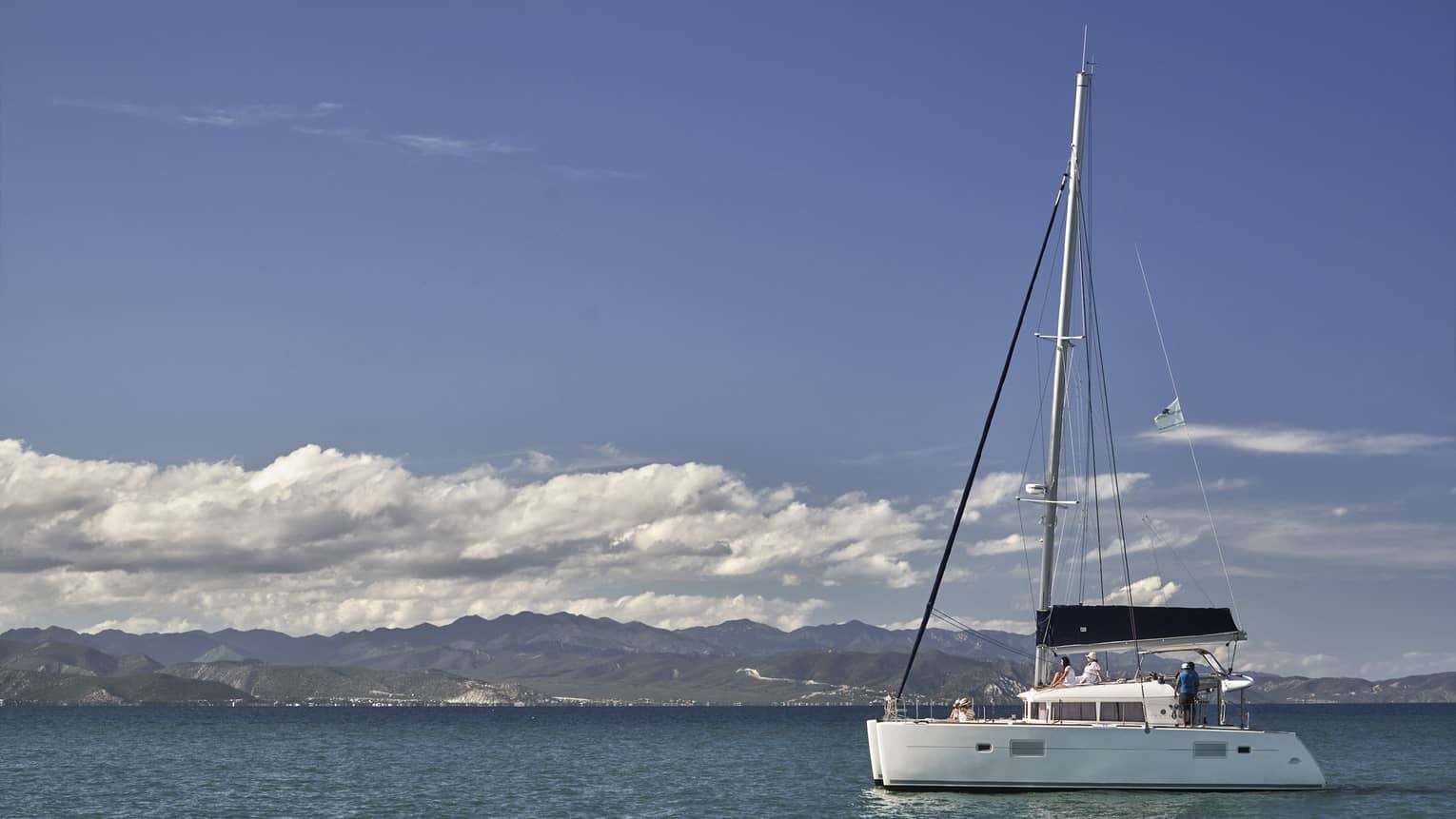 A catamaran on calm, blue water under a partly-cloudy sky with low green hills in the background and mountain ranges beyond.