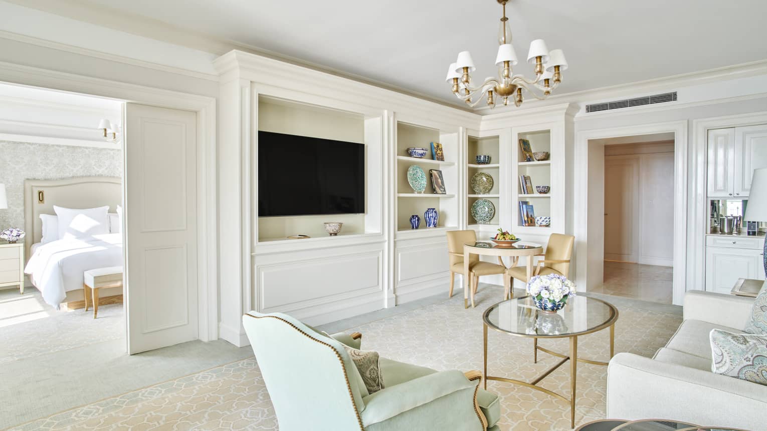 Suite living area with cream couch, round glass coffee table, flat-screen wall-mounted TV, doorway to bedroom