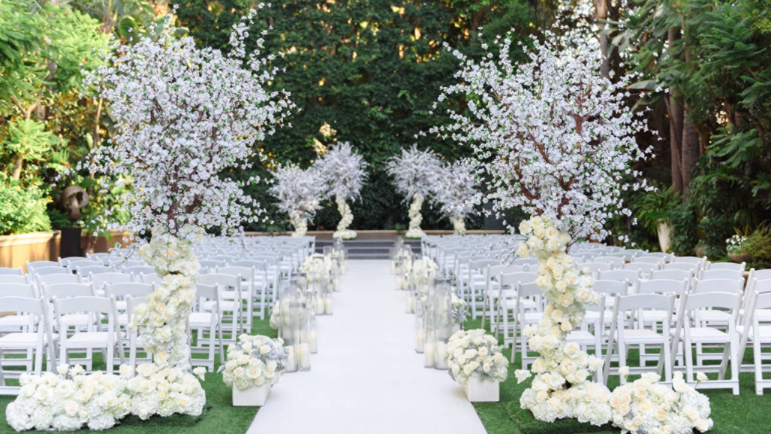 An outdoor wedding event space with rows of white chairs, white floral decor that includes flowering trees, and a white walkway leading to a raised platform in the background. The area is surrounded by trees and other greenery.