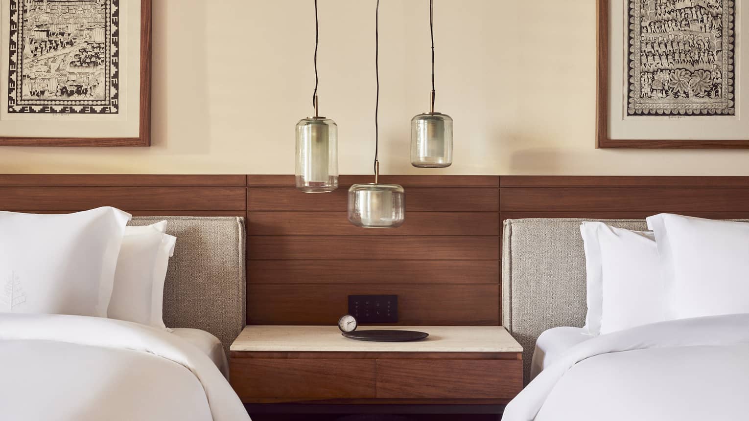 Cozy bedroom with two beds featuring white linens, a wooden headboard and a shared nightstand. Three pendant lights hang above, framed by artwork on the walls.