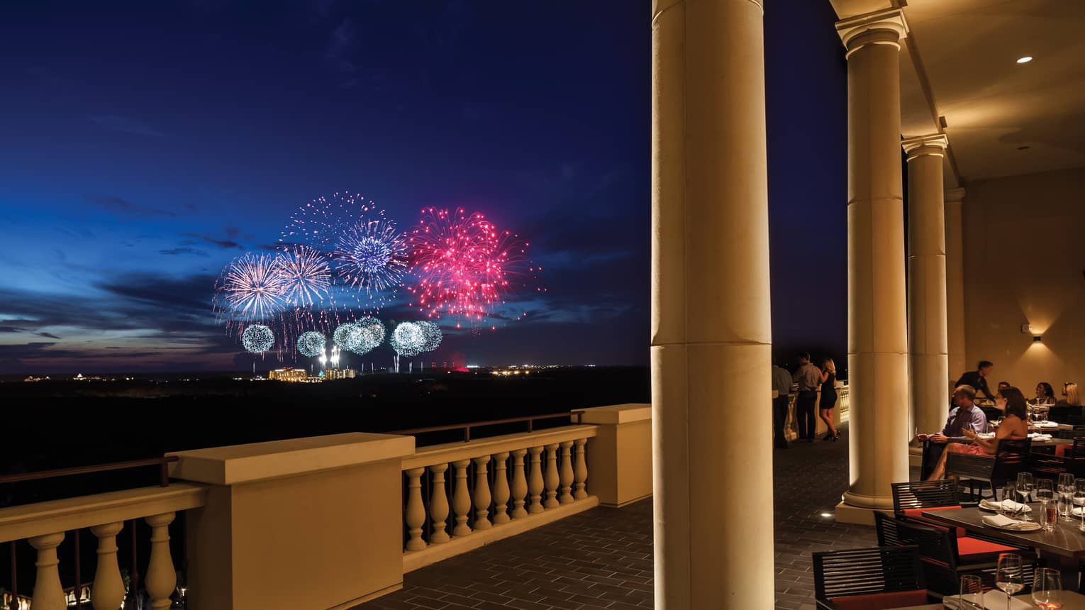 The terrace of a restaurant with tables and people watching fireworks in the distance.