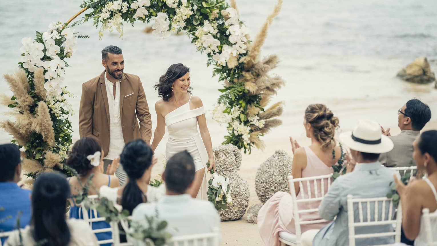 Bride and groom walk down aisle after ceremony on beach, flower arch and clapping guests