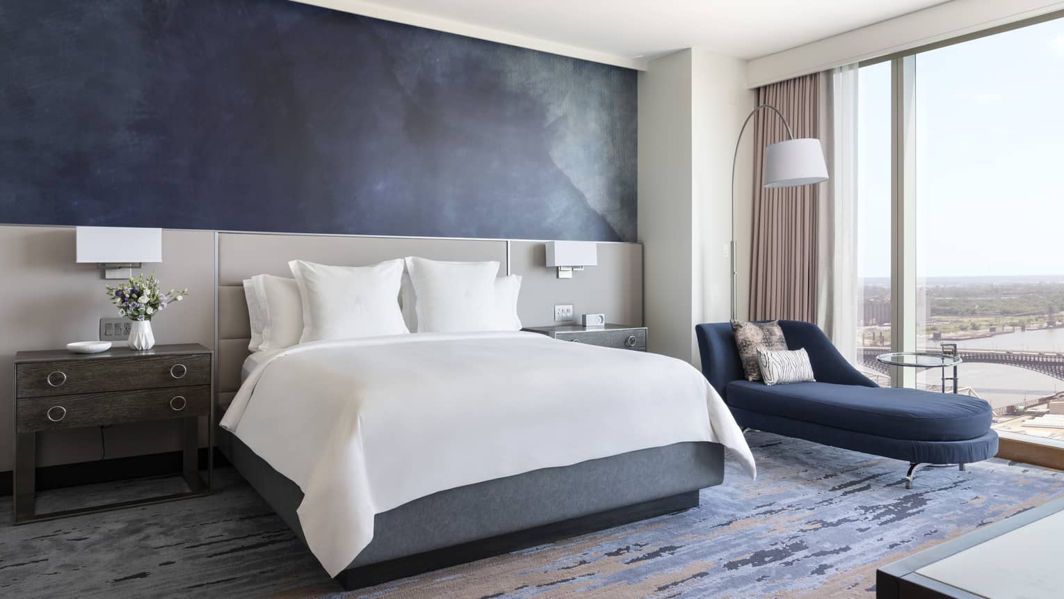 Bedroom with king bed, dark blue accent wall, dark blue chaise next to the window