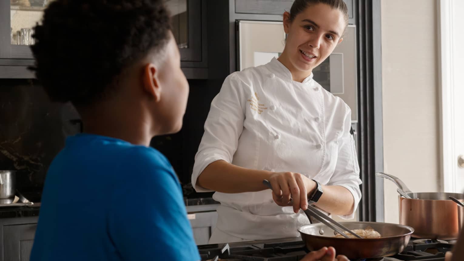 Rear close-up of a child looking on as a smiling chef prepares food in a copper frying pan, a copper pot on a nearby burner.