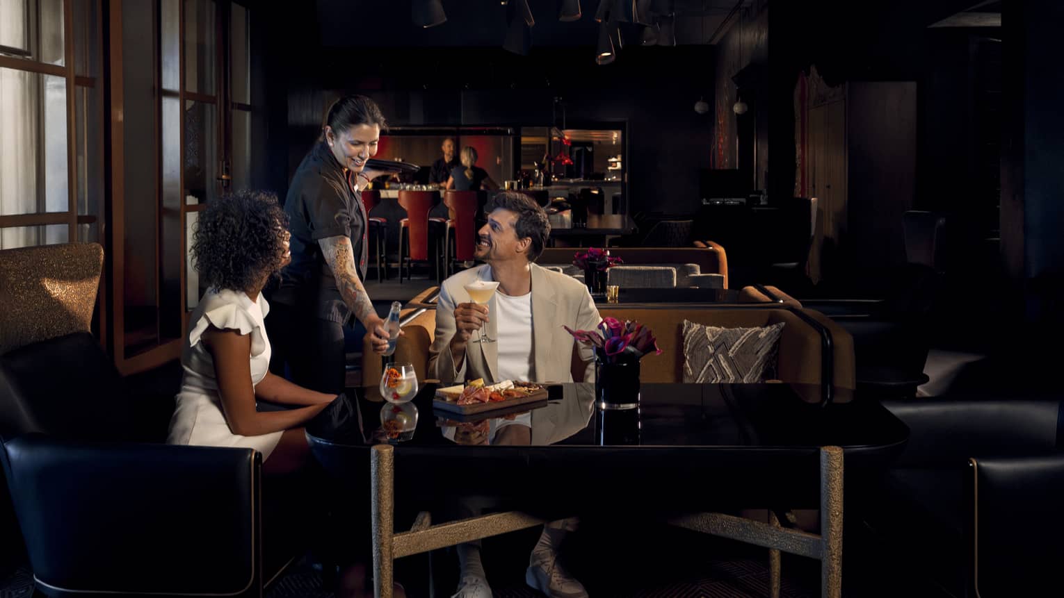 Two people wearing light-coloured cocktail attire sit on benches around a black table as a waitress brings them beverages