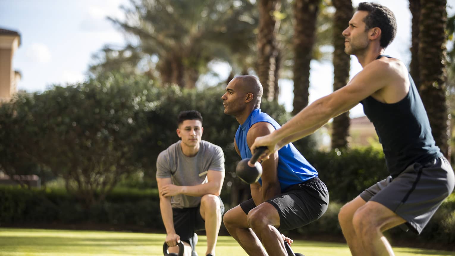 Four Seasons trainer watches as two men squat, lift kettlebell weights on lawn
