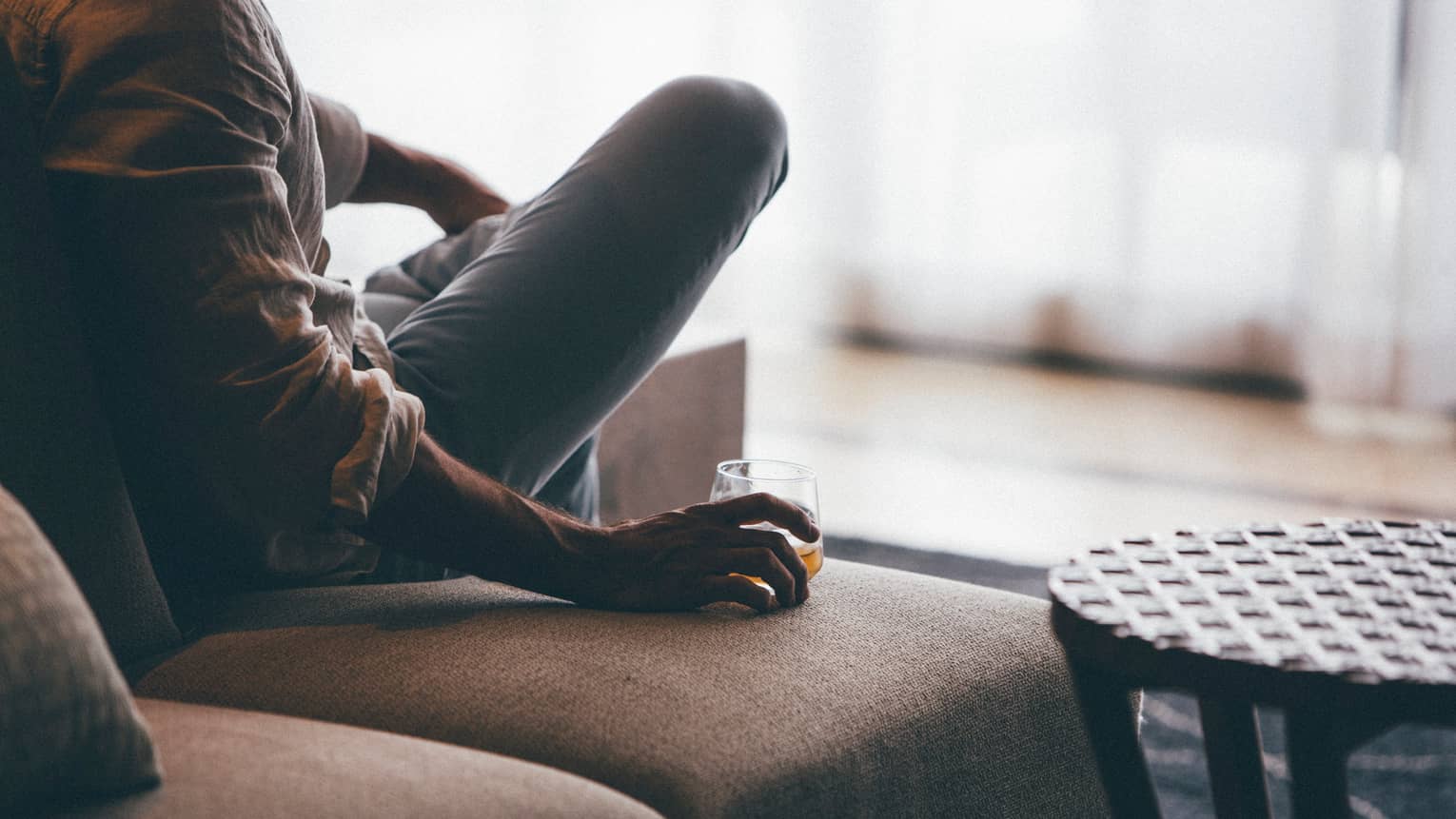 Close-up of man's arm with rolled-up dress shirt sleeves, blue jeans, holding glass of whisky on sofa