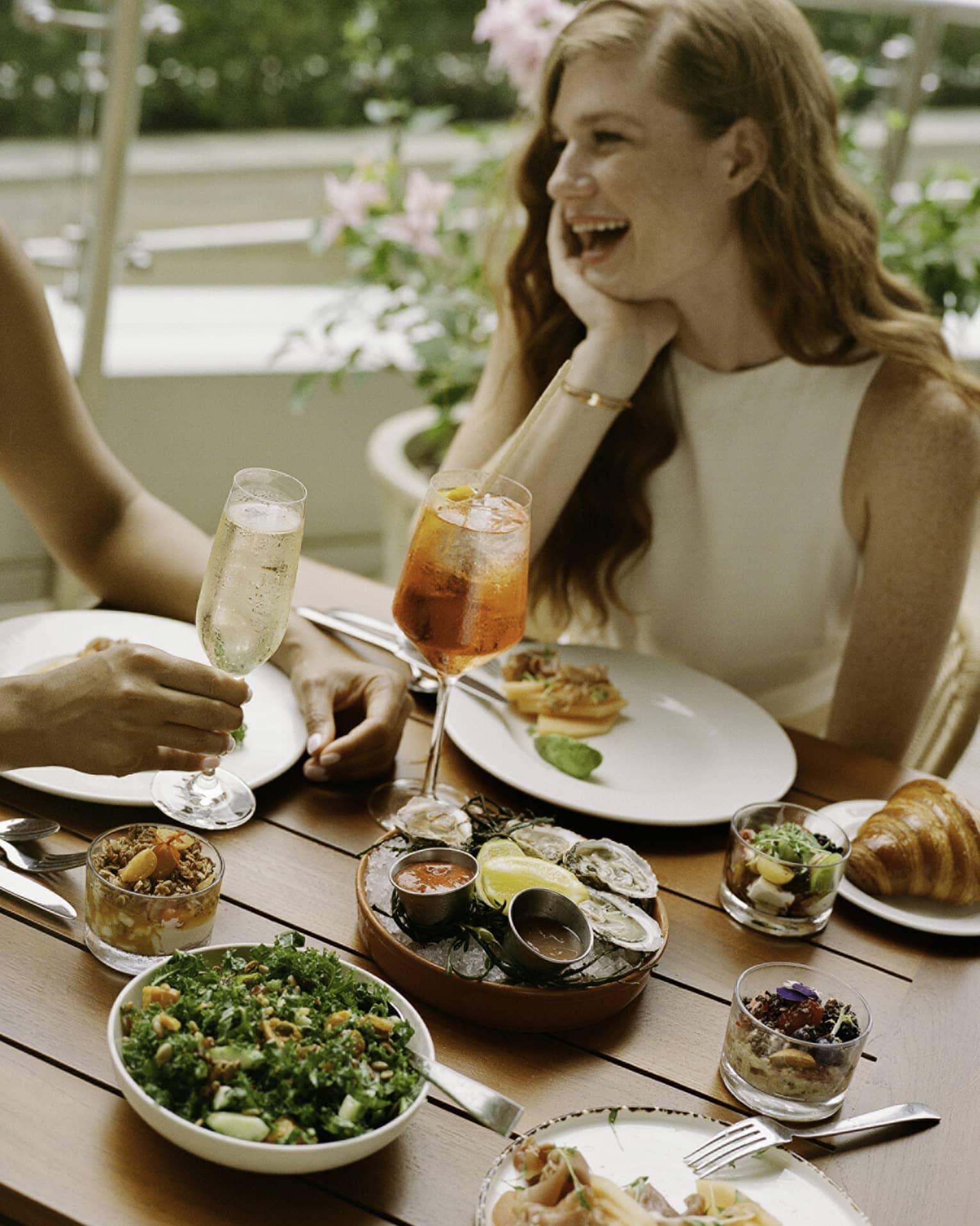 Laughing guests enjoying cold beverages, raw oysters with sauces, smoked salmon, a kale salad, muesli and croissants.