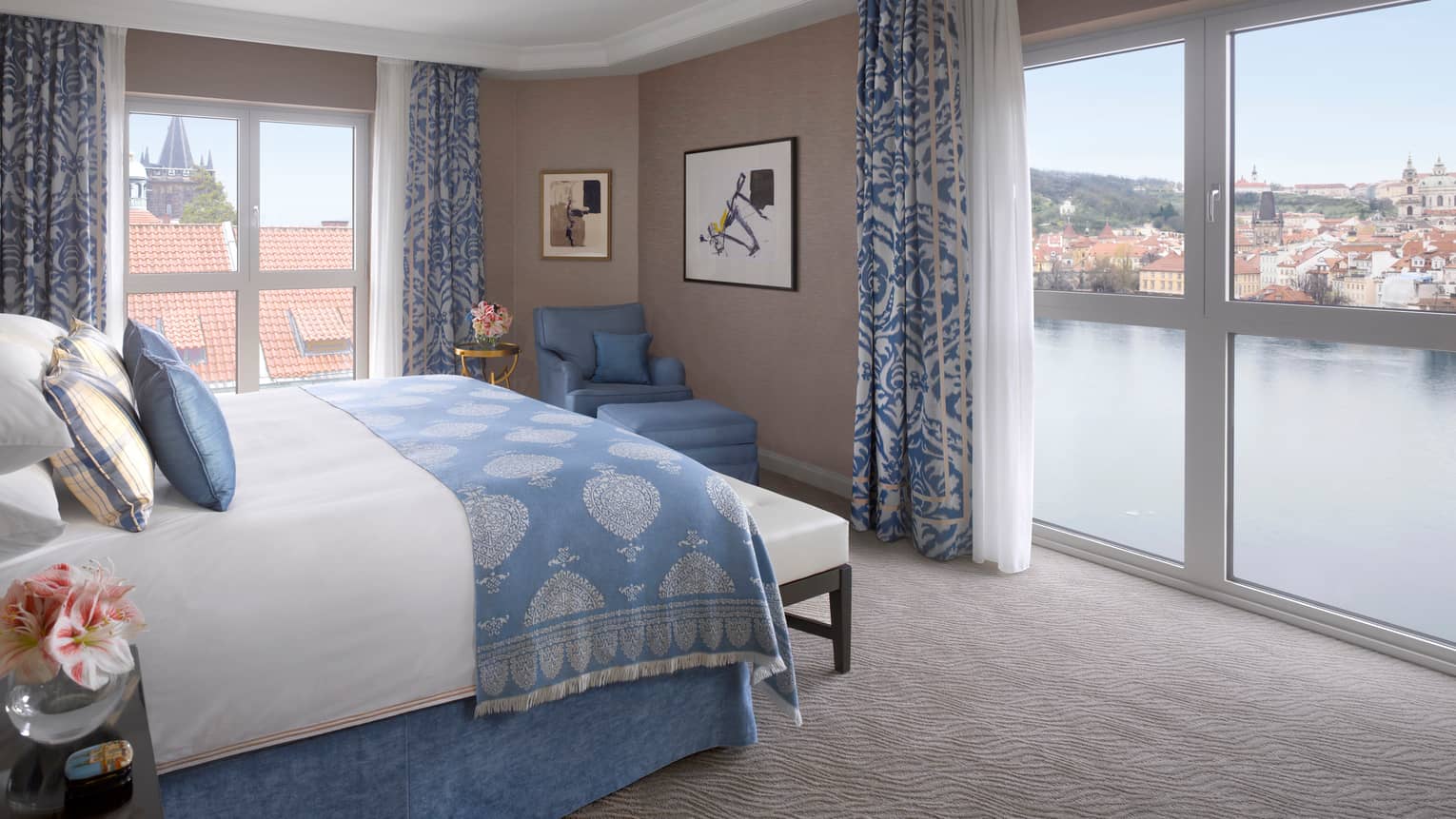 Premier Suite bed with blue blanket, pillow accents, in front of floor-to-ceiling corner windows overlooking Vltava River 