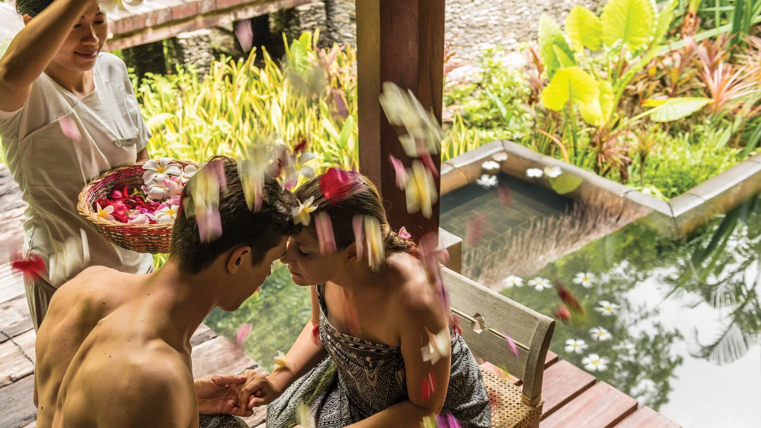 Couple on chairs, hold hands, foreheads together as woman sprinkles flower petals