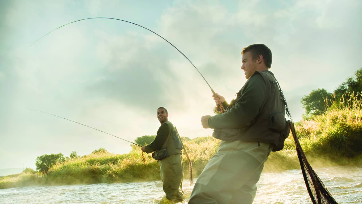 Two people fly fishing in a river, standing knee-deep in the water, surrounded by mist and greenery.