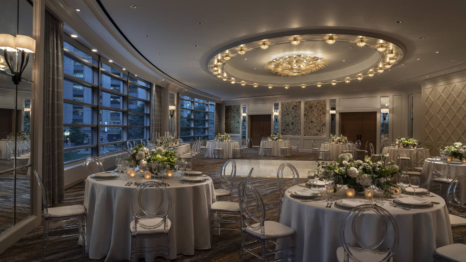 A dimly lit large meeting room with round tables covered in flowers and large windows.