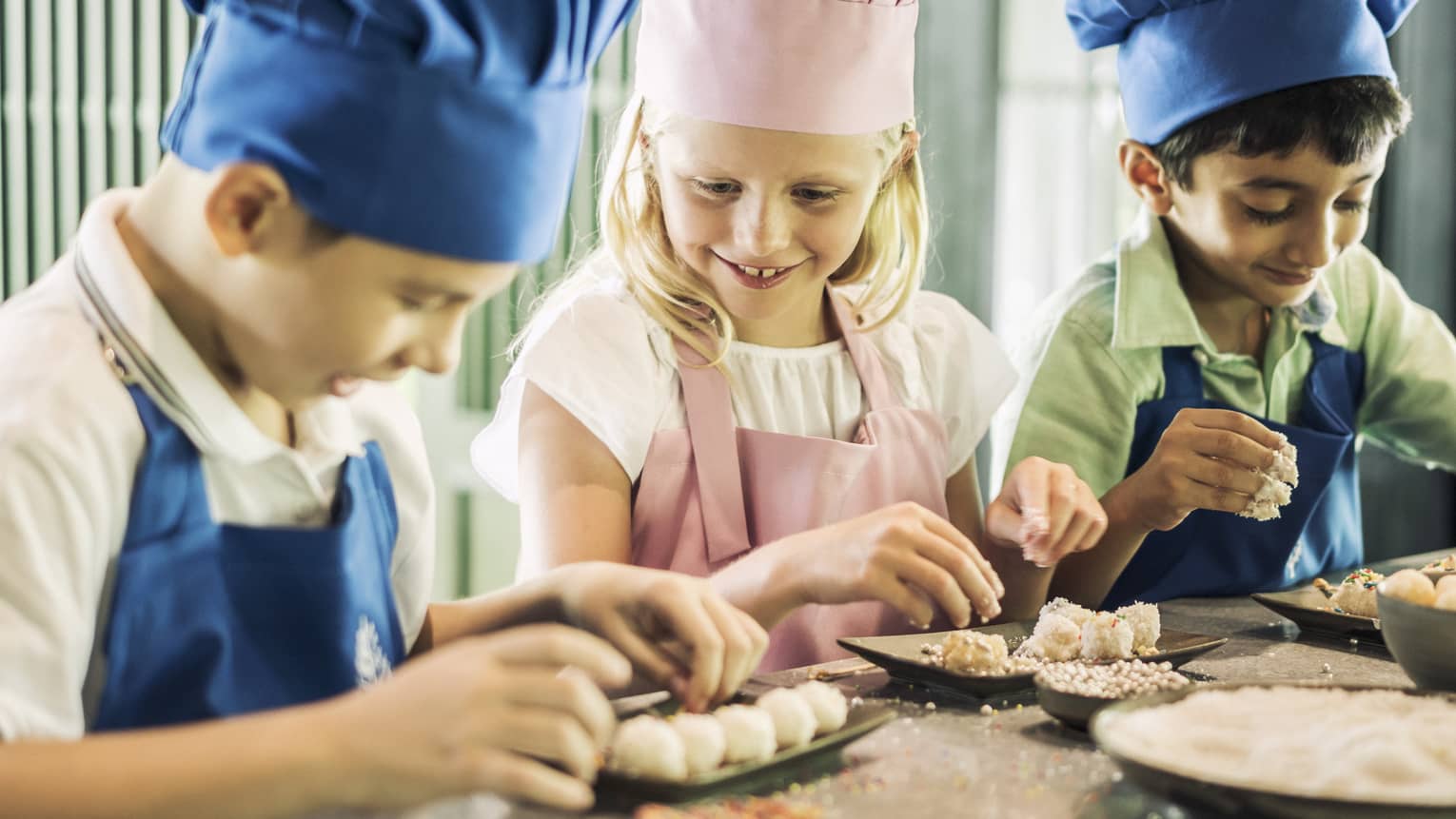 Three children in chef hats, aprons roll pastries at counter 