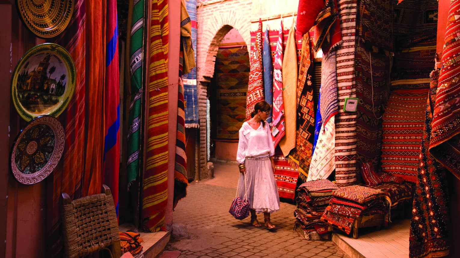 Woman holds purse, walks past hanging rugs, stacks of textiles in outdoor market 