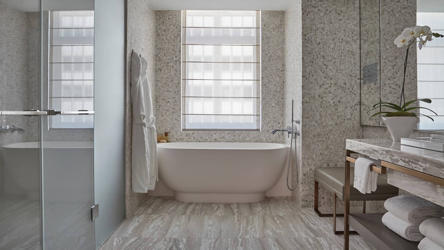 Elegant hotel bathroom with freestanding tub, mosaic tile walls, a vanity with an orchid, glass walk-in shower, and bathrobes hanging by a large window