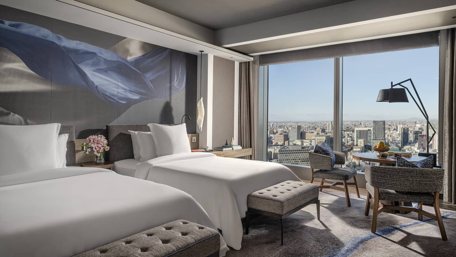 Guest room with two beds, upholstered benches and floor-to-ceiling windows offering panoramic city view, featuring modern décor